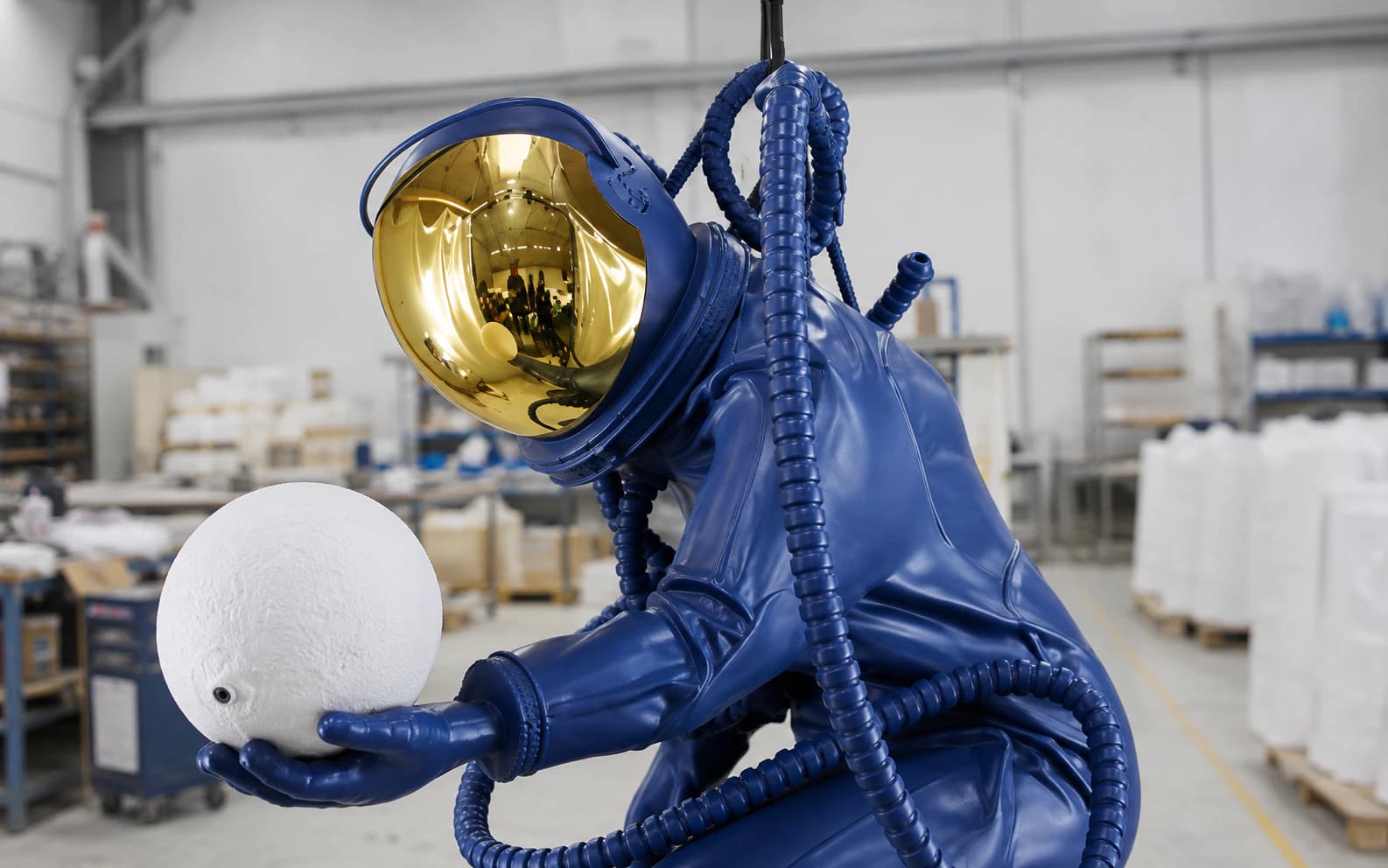 Close view of the astronaut sculpture showing the mirrored gold visor, suit folds, ribbed hoses, glove and white sphere.